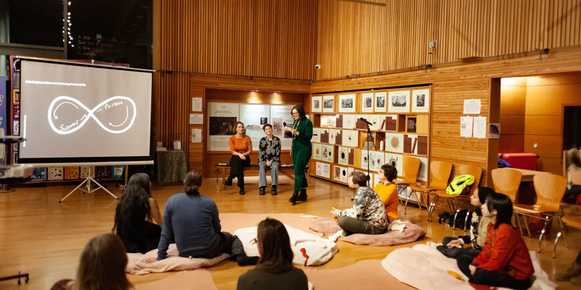 Joana talks to an audience, seated on cushions on the floor, with Amanda and Rose sat behind her.