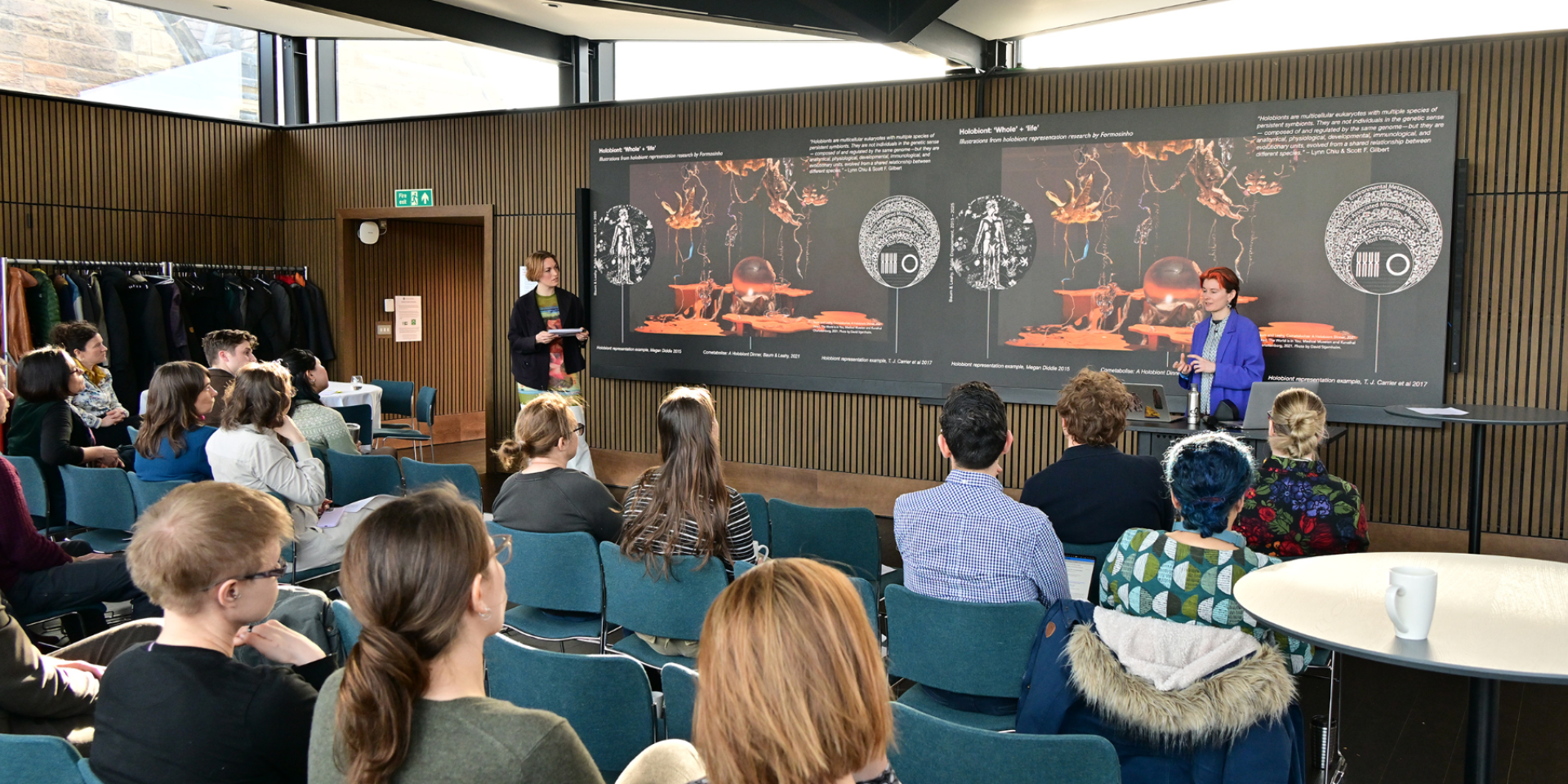 Baum and Leahy talk in front of a presentation of their art, with an audience in front.