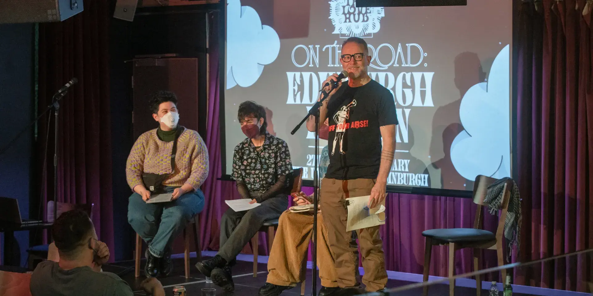 Will Nutland speaks on a small stage, with two guests sat behind him. 