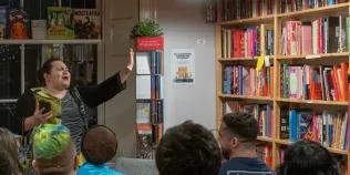 Georgia Bartlett-McNeil recites poetry to an audience with one arm raised and eyes shut, with bookshelves in the background.