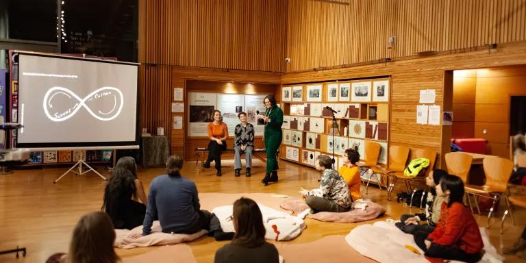 Joana talks to an audience, seated on cushions on the floor, with Amanda and Rose sat behind her.