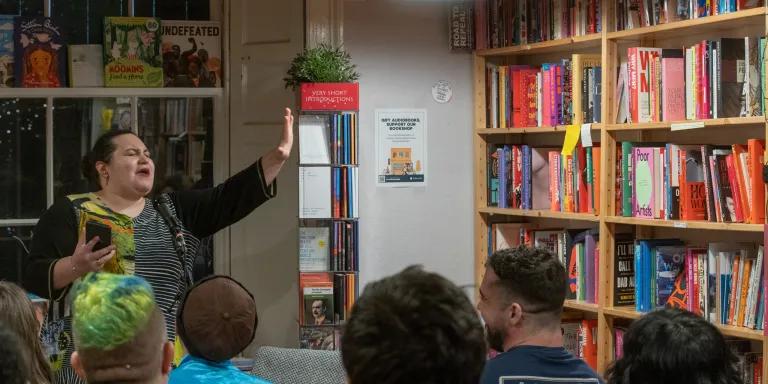 Georgia Bartlett-McNeil recites poetry to an audience with one arm raised and eyes shut, with bookshelves in the background.