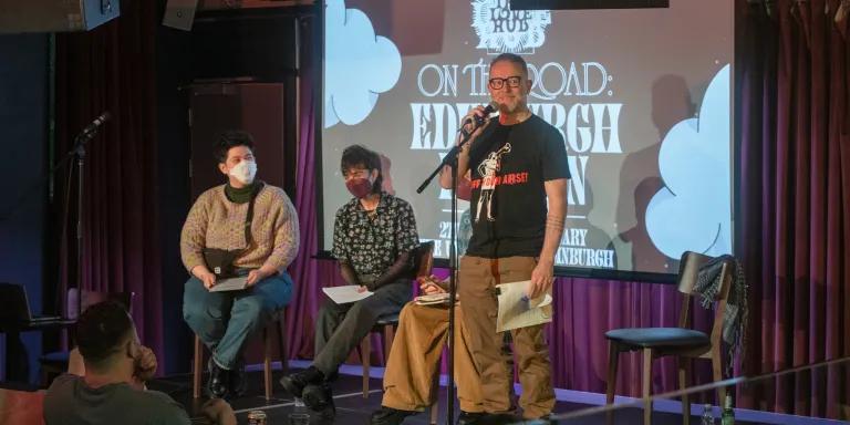 Will Nutland speaks on a small stage, with two guests sat behind him. 
