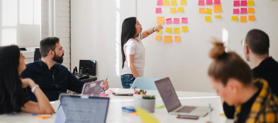 A group of people in a room looking at a colourful post it notes on a wall. They are sitting around a table with laptops and notebooks