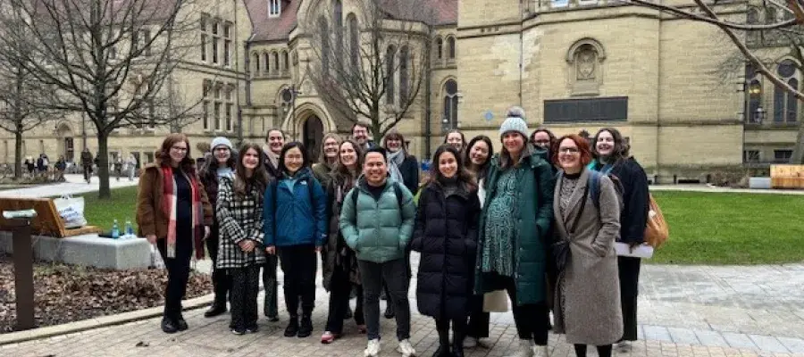 An image of a large group of people smiling and facing the camera, standing in front of the University of Manchester buildings.