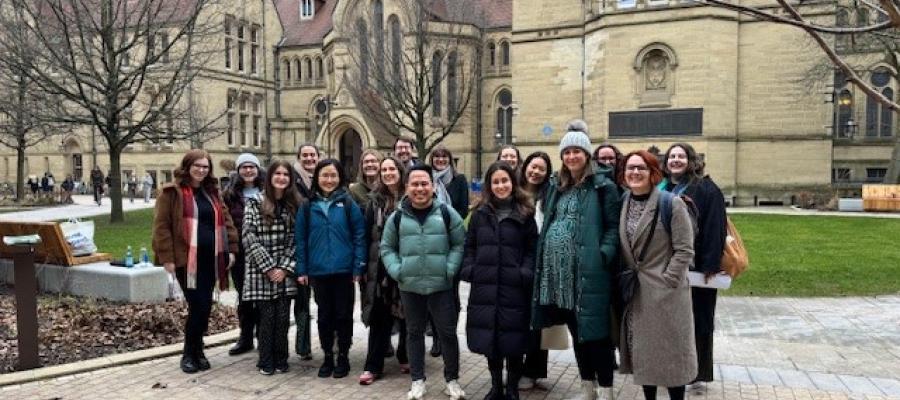 An image of a large group of people smiling and facing the camera, standing in front of the University of Manchester buildings.