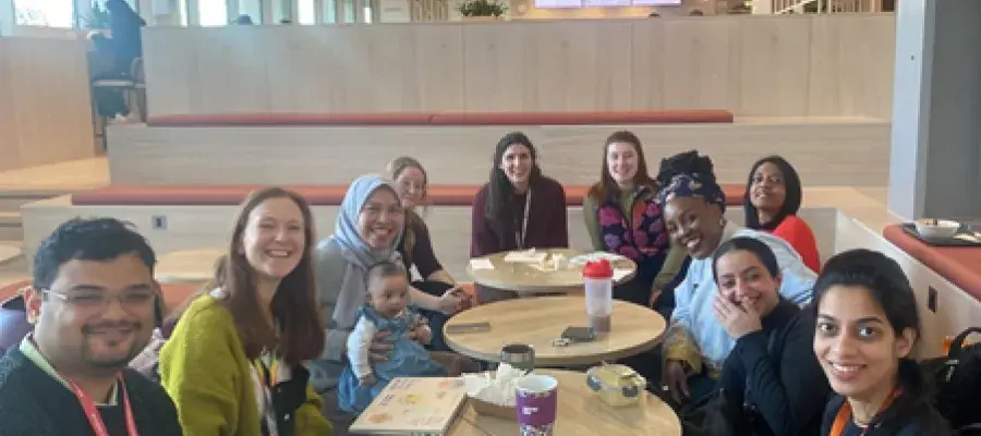 Photograph showing a group of PhD students sitting in the Usher Building, March 2025 (left to right: Shukanto Das, Eleanor Dixon, Fadila Wirawan and baby Asma Aisha Simatupang, Sophie Mackay, Katherine Edgley, Jazz Kirkwood, Thulani Ashcroft, Marylene Wamukoya, Areej Al-Mutairi, Durga Kulkarni)