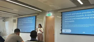 An image of a woman standing at the front of a room, in front of two projections where the text says 'Current and Future Priorities for UK Behavioural Research. A review of national and international strategy documents.'