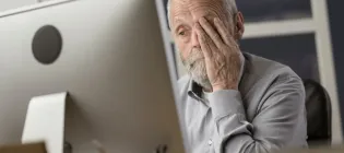 Older man looking tired in front of a computer, rubbing his eyes.