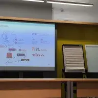 An image of a woman speaking in front of a projection, which shows a map of the UK with various University logos.