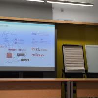 An image of a woman speaking in front of a projection, which shows a map of the UK with various University logos.