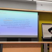 An image of a woman speaking in front of a projection, which has text on explaining today's aims. 