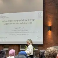 Professor Susan Michie standing in front of a whiteboard presentation, in front of an audience.