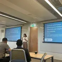 An image of a woman standing at the front of a room, in front of two projections where the text says 'Current and Future Priorities for UK Behavioural Research. A review of national and international strategy documents.'