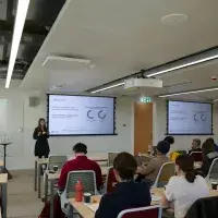 An image of a woman standing at the front of a busy room, speaking to the information presented on the 2 projection boards behind her.