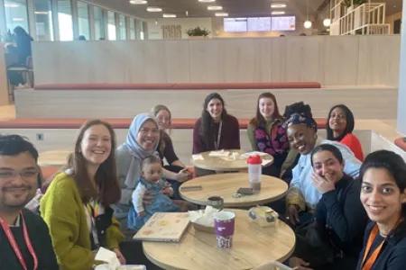 Photograph showing a group of PhD students sitting in the Usher Building, March 2025 (left to right: Shukanto Das, Eleanor Dixon, Fadila Wirawan and baby Asma Aisha Simatupang, Sophie Mackay, Katherine Edgley, Jazz Kirkwood, Thulani Ashcroft, Marylene Wamukoya, Areej Al-Mutairi, Durga Kulkarni)