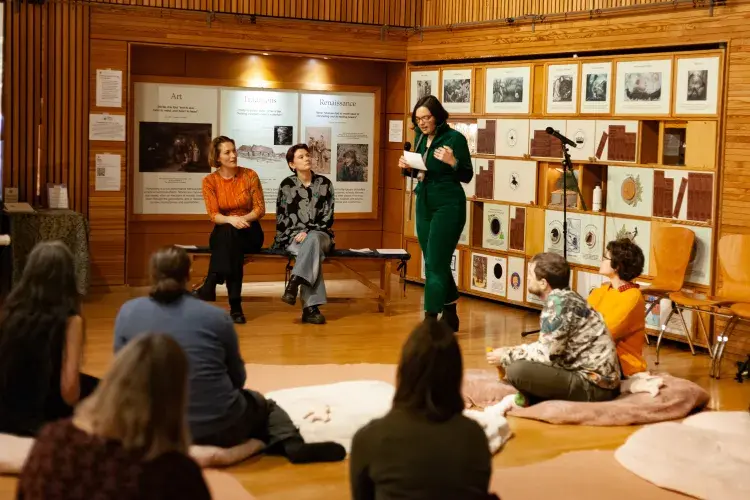 Joana talks to an audience, seated on cushions on the floor, with Amanda and Rose sat behind her.