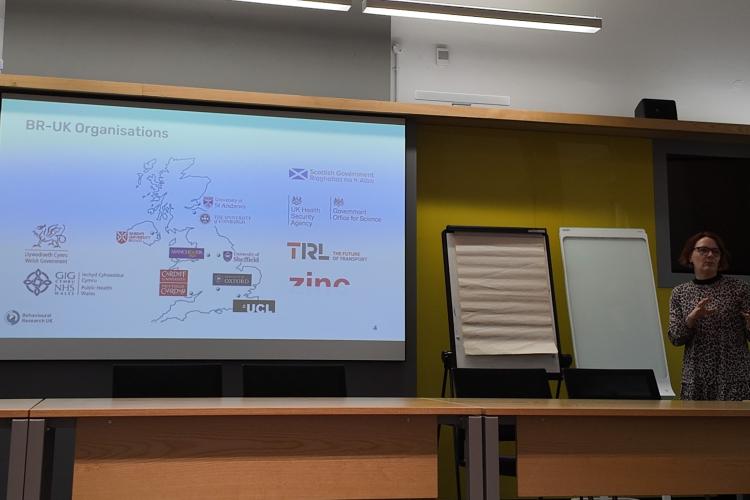 An image of a woman speaking in front of a projection, which shows a map of the UK with various University logos.