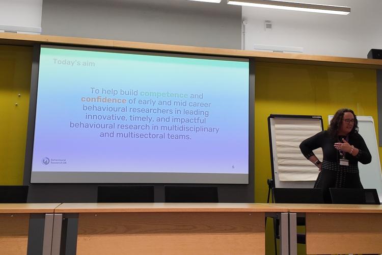 An image of a woman speaking in front of a projection, which has text on explaining today's aims. 