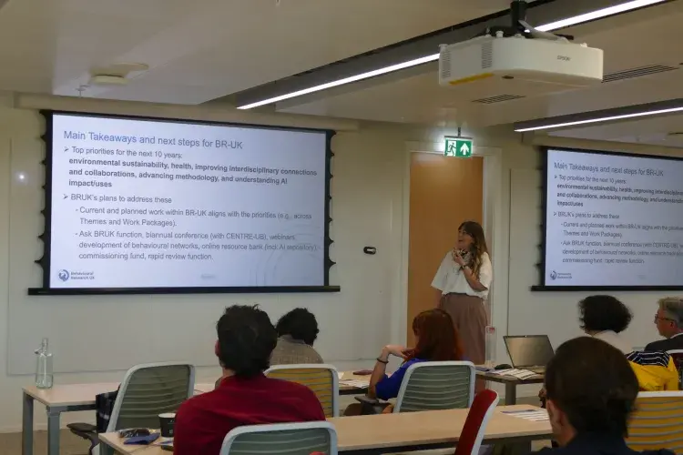 An image of a woman standing at the front of a room, in front of two projections where the text says  'main takeaways and next steps for BR-UK.'