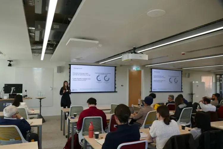 An image of a woman standing at the front of a busy room, speaking to the information presented on the 2 projection boards behind her.