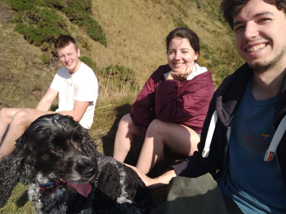 Three people sit outside with a dog. They are smiling at the camera