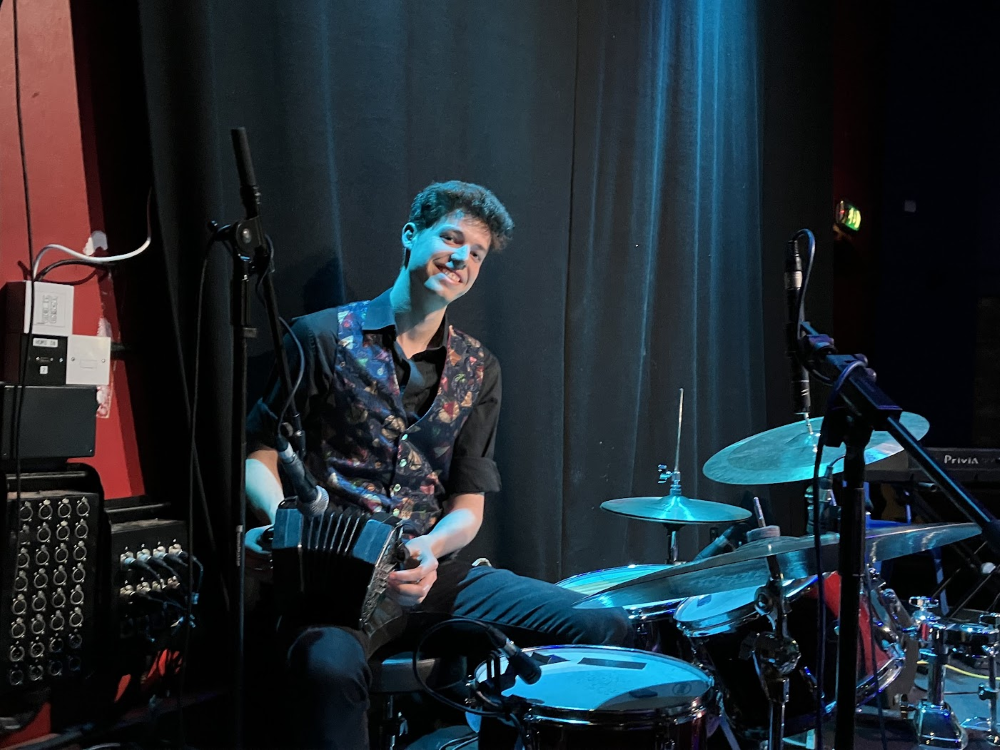 A young White man with curly brown hair sits at a drum set.