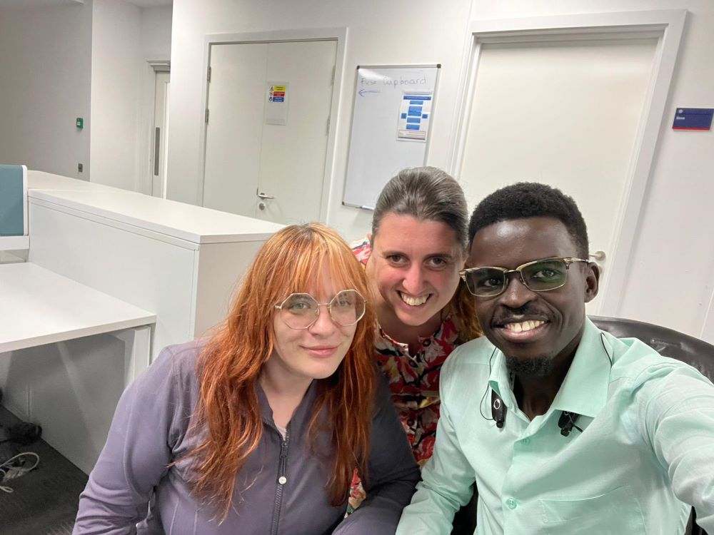 Three people are in the ACRC offices. The one on the left has red hair and glasses. The one in the middle has their arms around the two others. The one on the right is taking the photo and he wears a light green shirt and a wide smile.