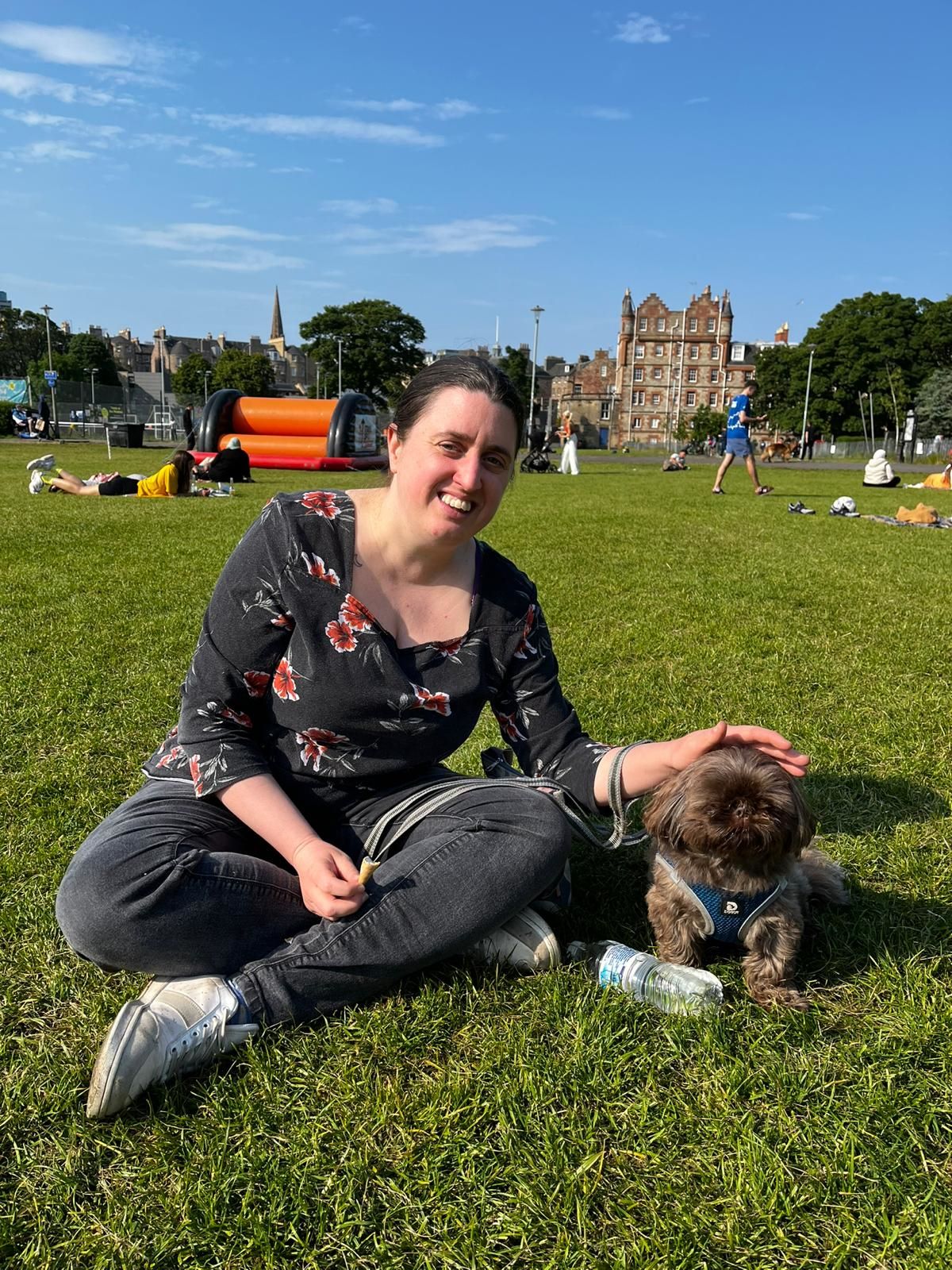 A smiling person with their hair back in a ponytail sits cross-legged on grass. There is a shit-tzu dog next to them.