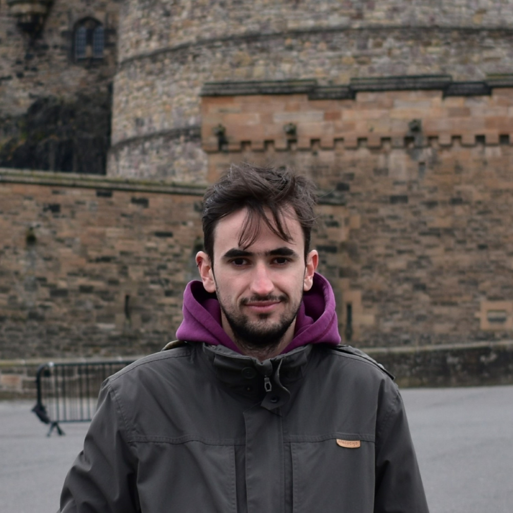 A young man with dark brown hair, a beard, and a jacket looks at the camera. A stone structure is behind him.