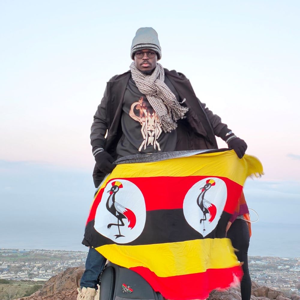 Wanok stands at the top of a creag, with the city of Edinburgh behind him. He holds the Ugandan flag.