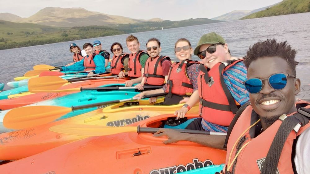 Nine people are lined up on a lake in kayaks. Wanok, a smiling young man with sunglasses, is holding the camera in selfie style.